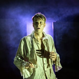 A young man stands looking confused and thoughtful, as though he has stopped in the middle of doing something. He wears a ragged off-white shirt with some of the buttons undone. The background is dark, but there is a doorway letting purple light and smoke into the space.