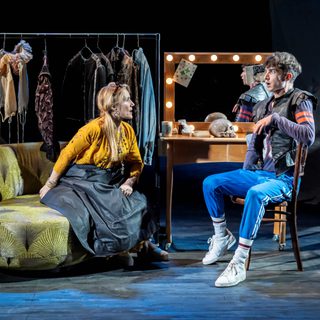 A man and a woman engage in a passionate conversion in a dressing room. The woman leans in, as if revealing something shocking, the man reacts as if shocked. There is a rail of costumes, dressing table and skull (presumably from Hamlet) behind them to set the scene.