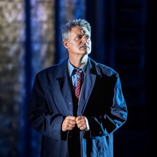 A middle-aged man looks into the distance with a contemplative expression. He wears a shirt, tie and long navy trench coat. Behind him is a blurry background of tall stained-glass windows.