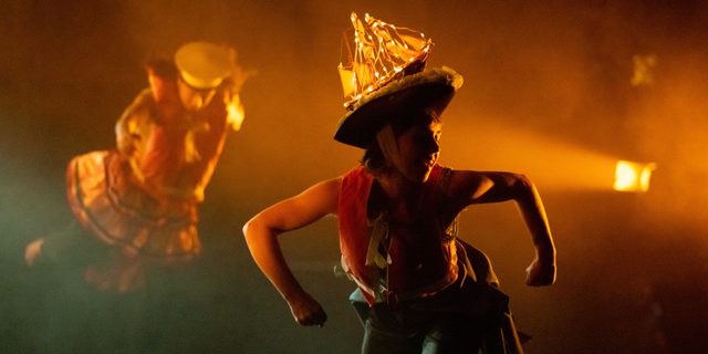 A dancer poses with their arms our at right angles and their fists clenched. They have a model ship attached to a hat they are wearing which has warm fairy lights draped on it. The lighting is a warm orange glow.