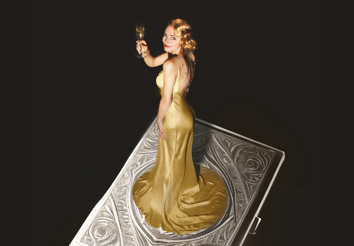 A giant, decorative, silver cigarette case floats against a black background. A young woman stands on top wearing a glamorous golden silk gown, holding a glass of champagne and looking to the camera with a smile. Her golden hair is curled and she wears red lipstick.