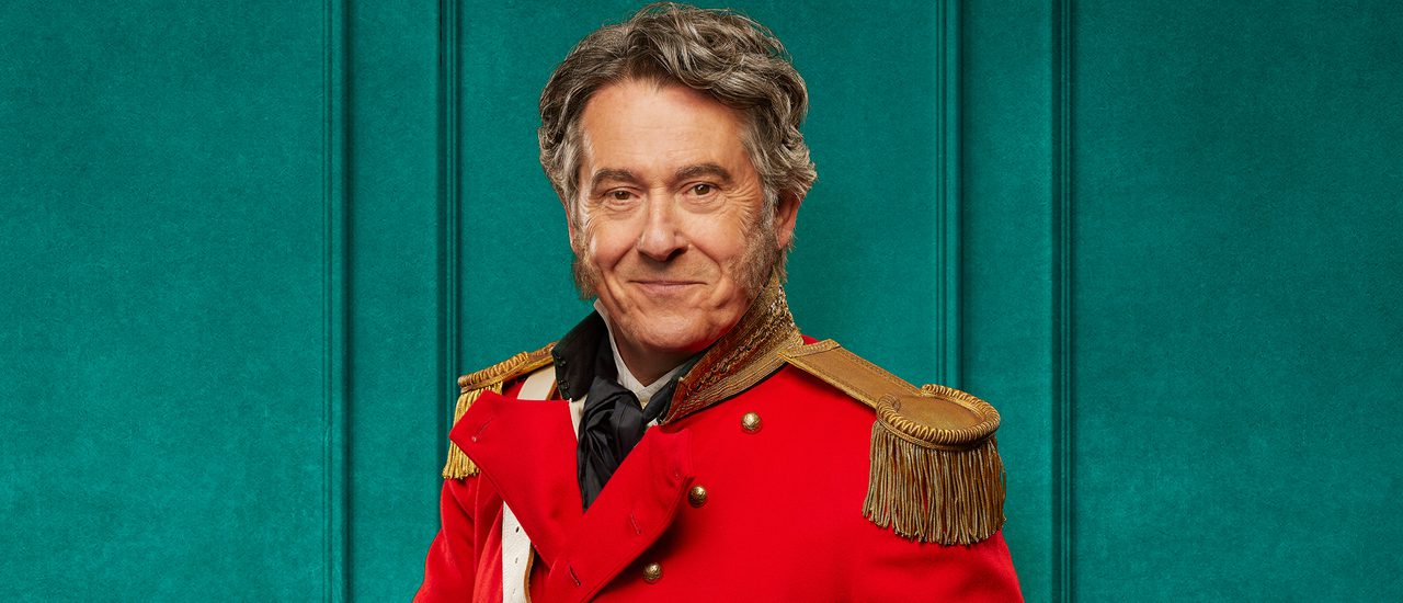 An older man with grey hair and exaggerated side burns stands proudly wearing ceremonial military full dress, including beige trousers and a red jacket with golden buttons, cuffs and epaulettes. He holds a glass of whiskey and looks straight down the camera with a knowing smile.