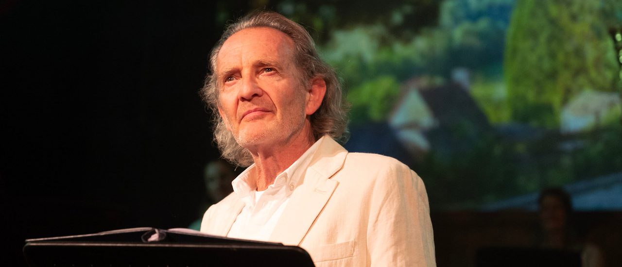Anton Lesser, an older man with long grey hair, stands behind a lectern, wistfully looking out.