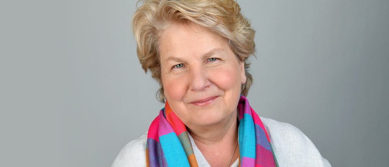Sandi Toksvig, a woman with short blonde hair, smiles warmly, wearing a white shirt and colourful pink, purple and blue checked scarf.