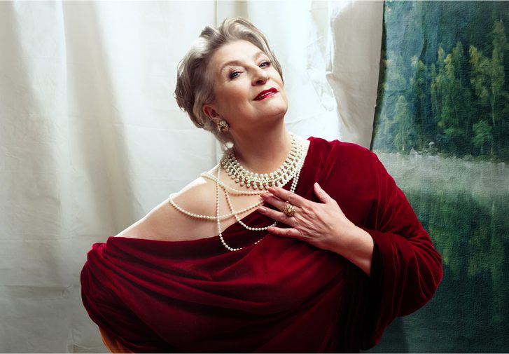 An older woman leans to one side, endearingly looking at the camera with a slight smile. She is heavily made up with dark red lipstick and has her grey hair tied back. She wears a luxurious dark red velvet one-shoulder dress, layers of pearls, a large ring and big round earrings.