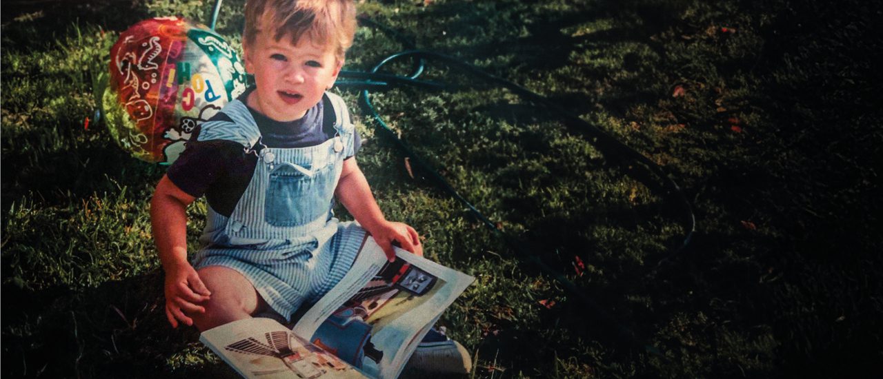 A vintage photograph of a young boy sitting in the grass, holding a magazine with a colourful beach ball behind him. He wears a dark blue t-shirt and blue and white stripey dungarees.