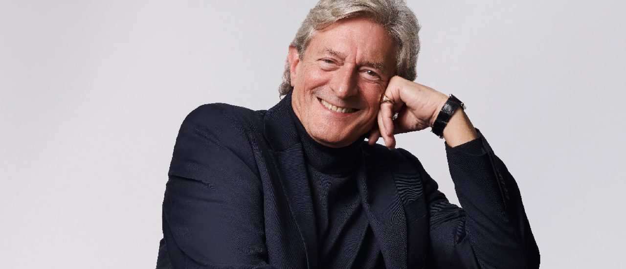 Nigel Havers, an older man with grey hair, leans on one hand and smiles brightly. He wears a dark polo neck, blazer and watch.
