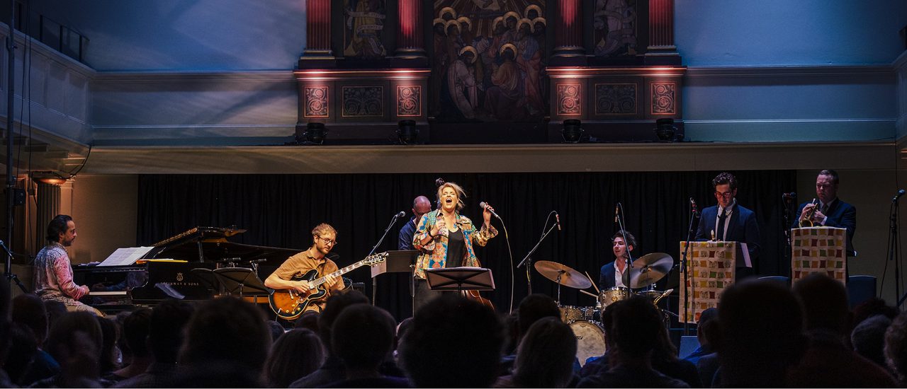 Clare Teal, a woman with blonde hair, wearing a colourful patterned shirt and black top, sings passionately, eyes closed, holding a microphone, with a music stand in front of her.