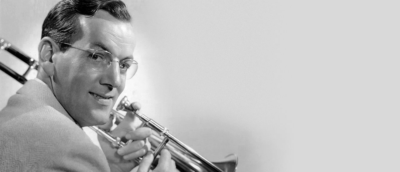 A black-and-white photograph of a man with a vintage comb over holding a trumpet, looking back over one shoulder and smiling warmly.
