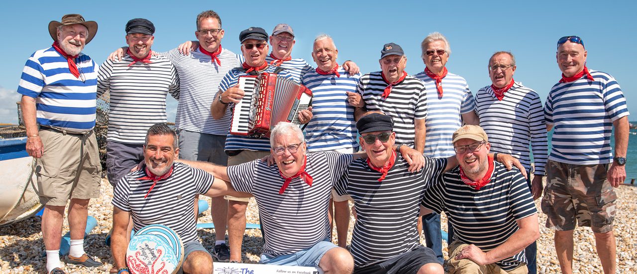 Three men wearing white and blue stripey tops with red neckerchiefs. One plays an accordion whilst another looks out to the side, open mouthed, exclaiming happily.