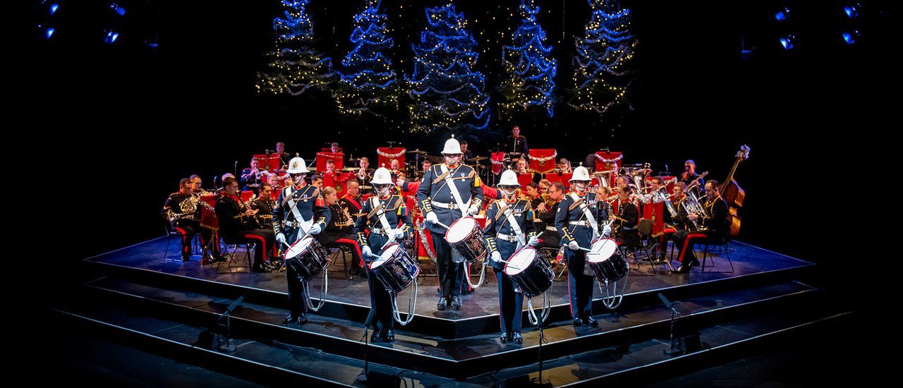 The Band of HM Royal Marines stand on the Festival Theatre stage dressed in military full dress, facing out front. Five drummers stand at the front, with the rest of the band seated behind. Behind them are five blue-lit Christmas Trees.