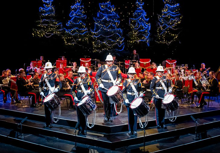 The Band of HM Royal Marines stand on the Festival Theatre stage dressed in military full dress, facing out front. Five drummers stand at the front, with the rest of the band seated behind. Behind them are five blue-lit Christmas Trees.
