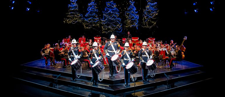 The Band of HM Royal Marines stand on the Festival Theatre stage dressed in military full dress, facing out front. Five drummers stand at the front, with the rest of the band seated behind. Behind them are five blue-lit Christmas Trees.