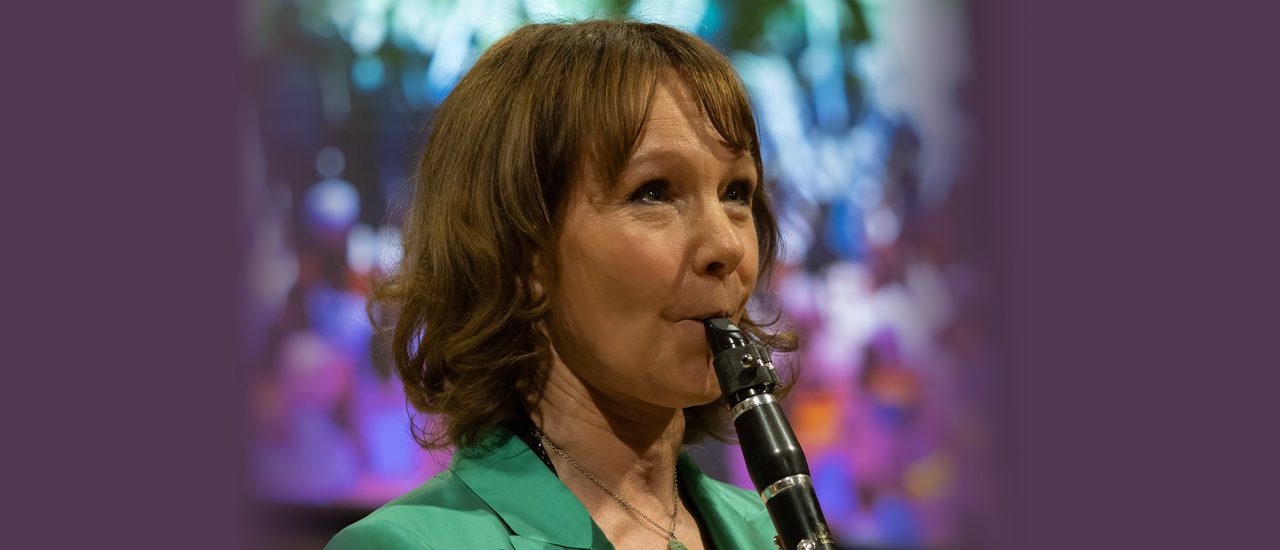 Emma Johnson MBE, a woman with mid length brown hair wearing a green blazer, plays a clarinet. In the background is a colourful graphic projection with shades of blue, purple and green.