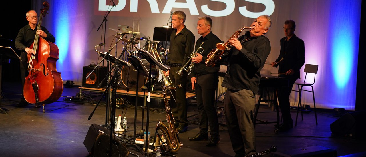 Three musicians side on playing various brass instruments, wearing matching black shirts. In the background, white text reads 'Shear Brass'.