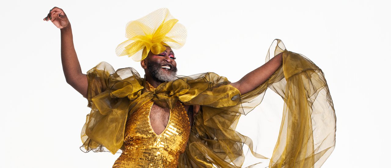 A Black drag performer with a big grey beard wears a dazzling, gold, sequined ball gown with puffy tulle sleeves, an exaggerated train and a yellow flower headpiece. They smile and hold both arms out in a sassy flourish.