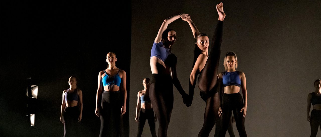 Dancers are on a simple black stage. At the front of the image, two dancers link hands as the one on the right kicks her leg high into the air. The dancers behind them are stood still with an engaged energy.