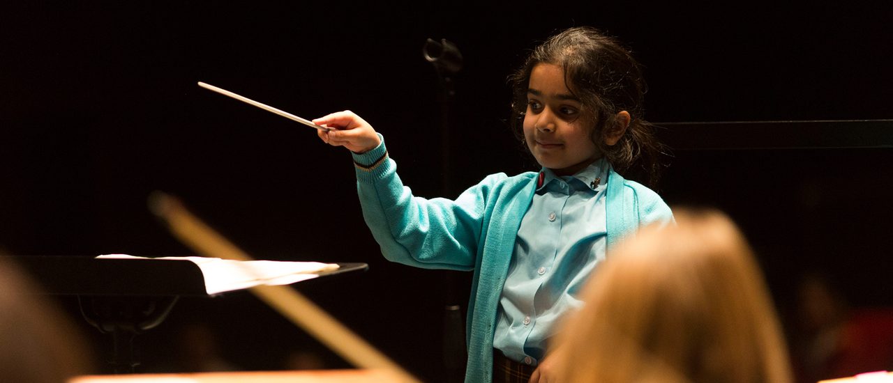 A young girl is smiling as she waves a conductor's baton at an orchestra. In front of her is a music stand. She wears a blue school uniform.