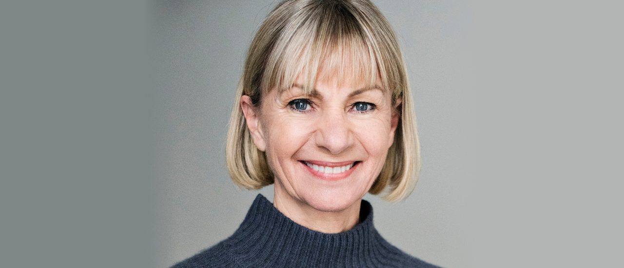 Kate Mosse, a woman with a neat blonde bob, smiles warmly. She wears a dark grey, turtle-neck jumper.