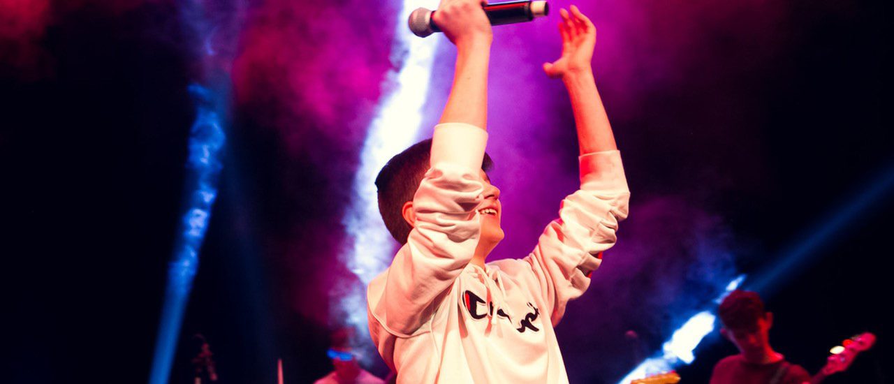 A boy in a white sweatshirt holds his arms above his head, holding a microphone. Several spotlit musicians are behind him.