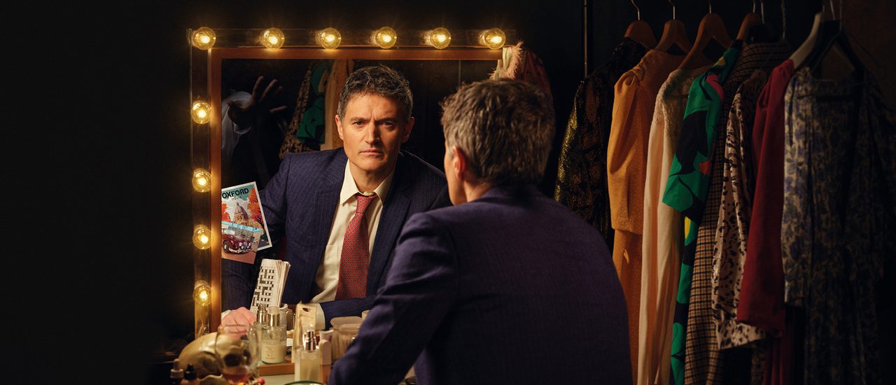 A middle-aged man with short, grey hair sits in front of a dressing table, looking into a mirror surrounded by lights. He is photographed from behind, revealing his reflection. He has an intense stare and a frown. He wears a blue pin -stripe suit jacket, white shirt and red patterned tie and holds a folded newspaper. The dressing table is cluttered with an array of objects, including a nearly-empty wine glass, skull, make-up, pills and toiletries.