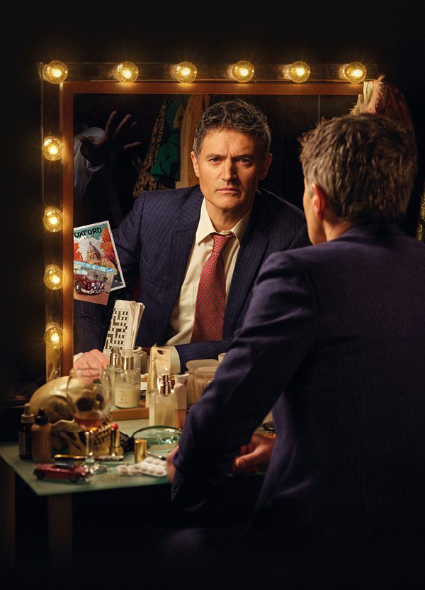 A middle-aged man with short, grey hair sits in front of a dressing table, looking into a mirror surrounded by lights. He is photographed from behind, revealing his reflection. He has an intense stare and a frown. He wears a blue pin -stripe suit jacket, white shirt and red patterned tie and holds a folded newspaper. The dressing table is cluttered with an array of objects, including a nearly-empty wine glass, skull, make-up, pills and toiletries.