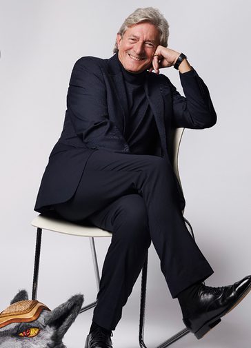 Nigel Havers, an older man with grey hair, leans on one hand and smiles brightly. He wears a dark polo neck, blazer and watch.