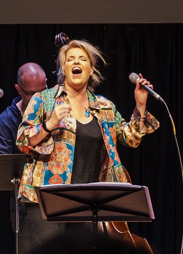Clare Teal, a woman with blonde hair, wearing a colourful patterned shirt and black top, sings passionately, eyes closed, holding a microphone, with a music stand in front of her.