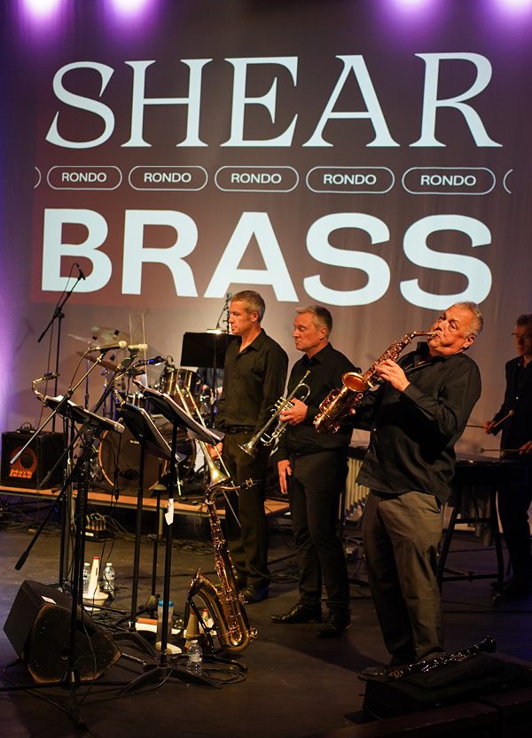Three musicians side on playing various brass instruments, wearing matching black shirts. In the background, white text reads 'Shear Brass'.