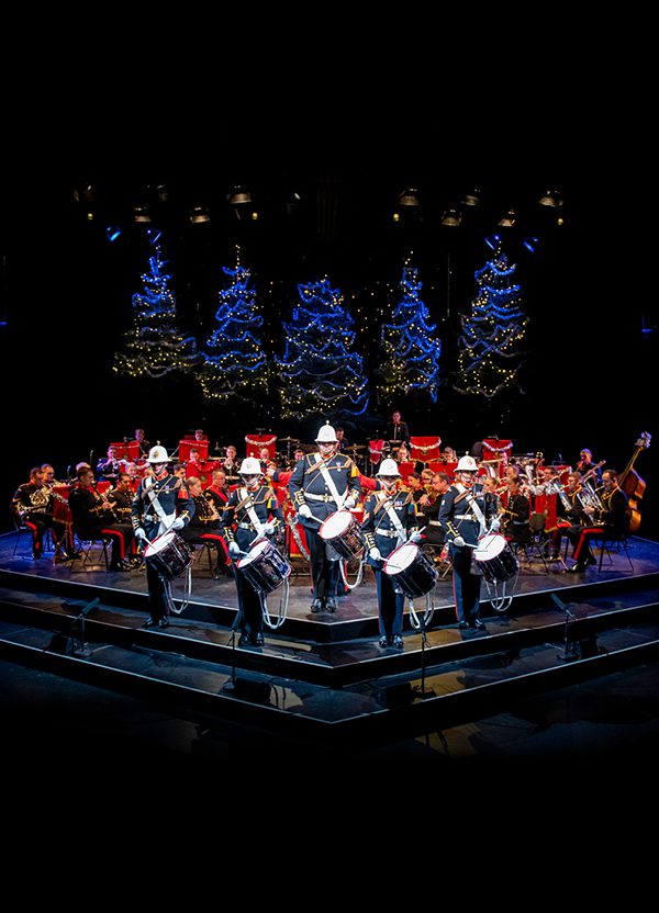 The Band of HM Royal Marines stand on the Festival Theatre stage dressed in military full dress, facing out front. Five drummers stand at the front, with the rest of the band seated behind. Behind them are five blue-lit Christmas Trees.