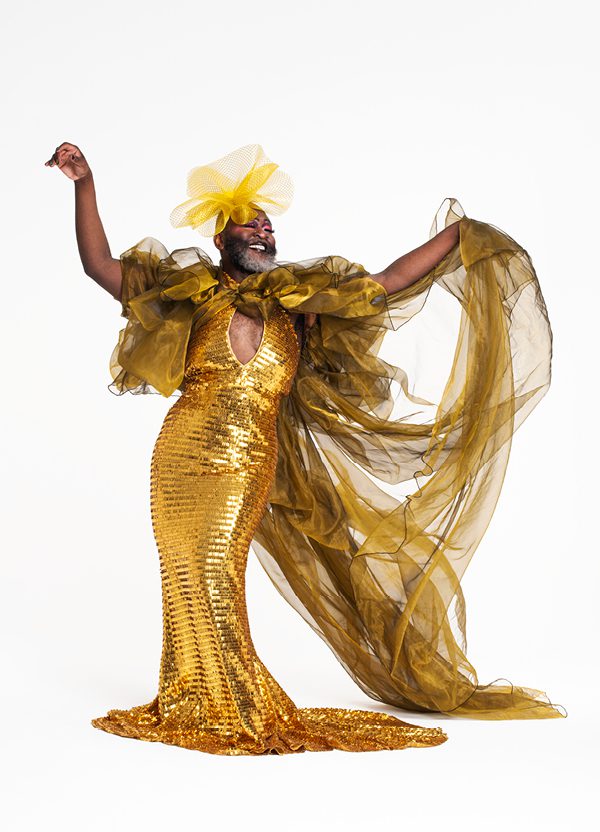 A Black drag performer with a big grey beard wears a dazzling, gold, sequined ball gown with puffy tulle sleeves, an exaggerated train and a yellow flower headpiece. They smile and hold both arms out in a sassy flourish.