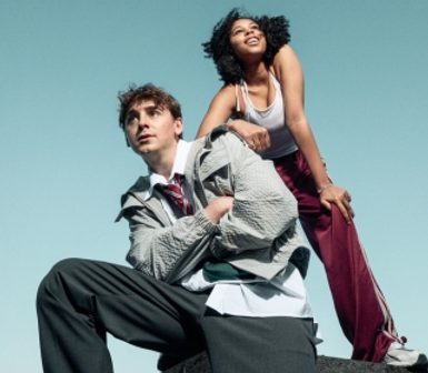 Two young performers stood leaning against each other and looking in the distance, behind them is a blue sky.