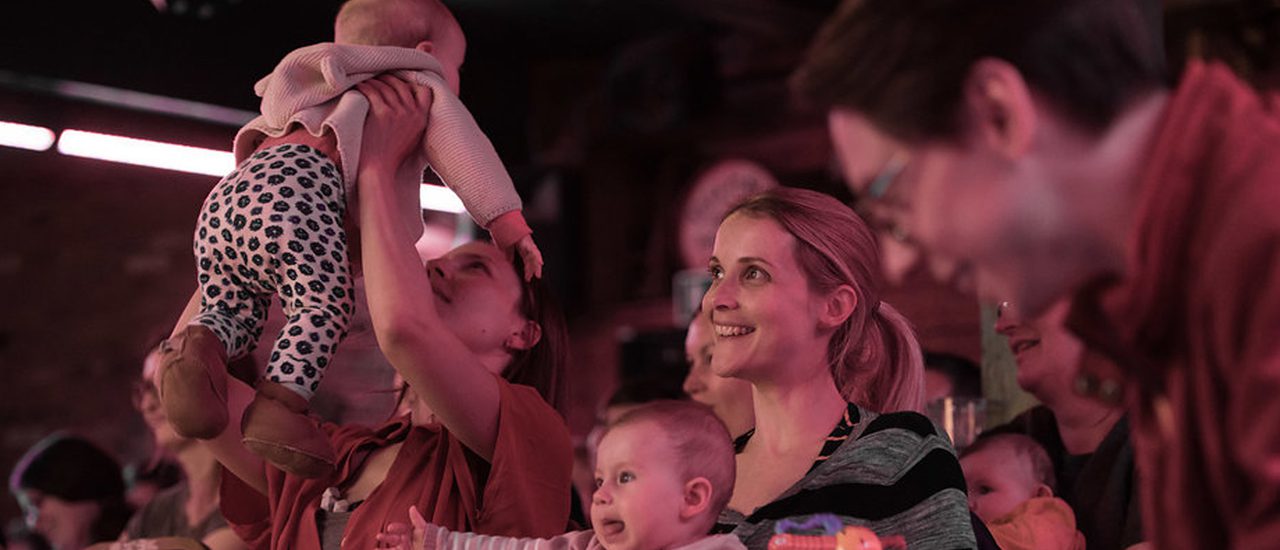 A young woman with a lively baby on her lap sits laughing. She's surrounded by other men and women with babies; on the right, another woman holds her baby up in the air.