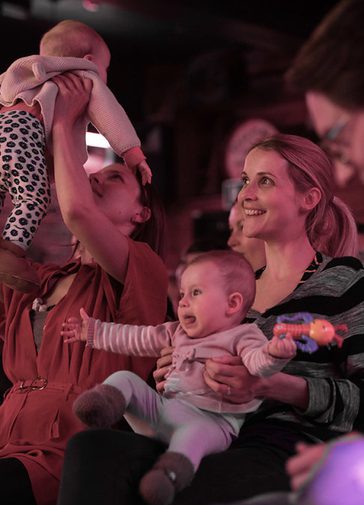 A young woman with a lively baby on her lap sits laughing. She's surrounded by other men and women with babies; on the right, another woman holds her baby up in the air.
