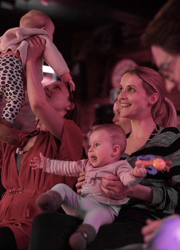 A young woman with a lively baby on her lap sits laughing. She's surrounded by other men and women with babies; on the right, another woman holds her baby up in the air.
