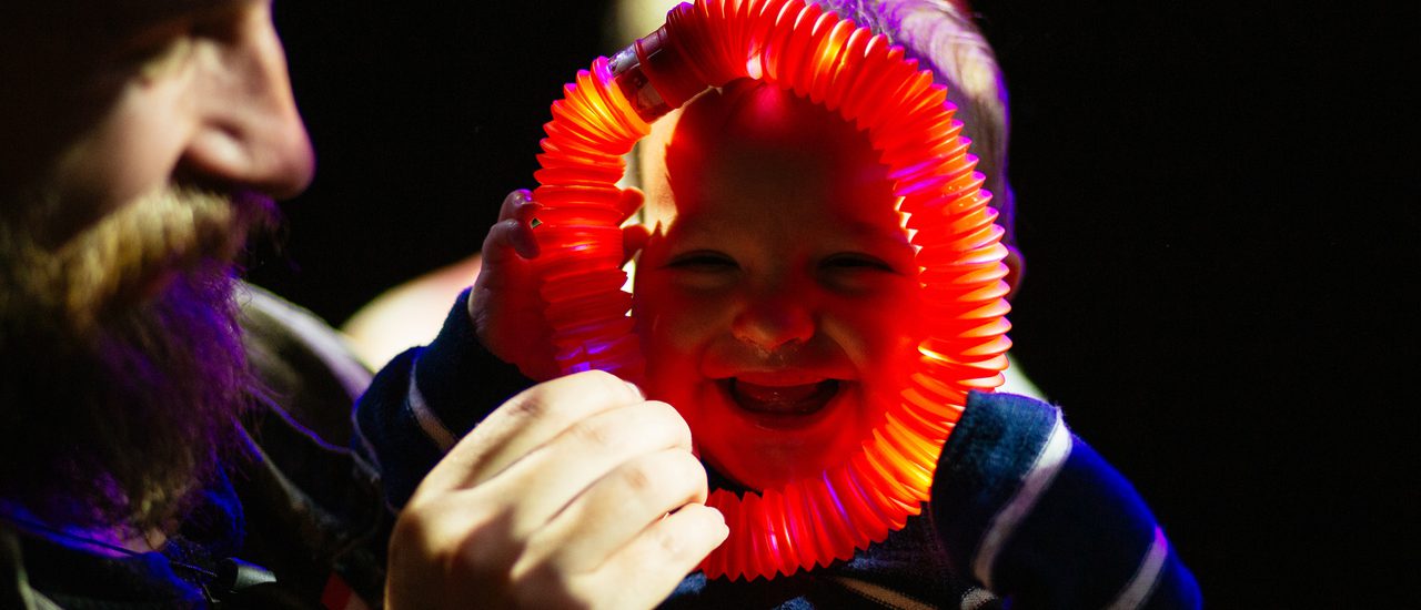 A bearded man holds an illuminated red light ring up to a laughing baby's face.