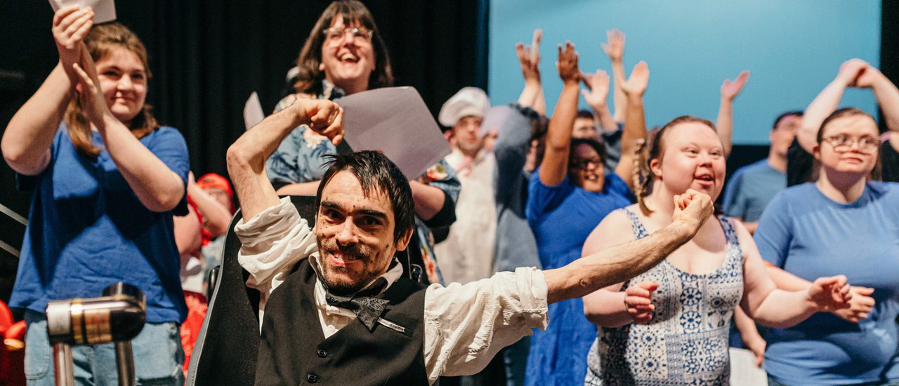 A group of learning-disabled and neuro-divergent people performing, dancing and laughing. In the forefront, a male wheelchair user, wearing a white shirt and black waistcoat, enthusiastically waves a piece of paper above his head, his other arm spread out wide.