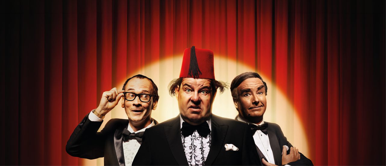 Three men stand together in front of a red curtain, lit with a golden spotlight. They all wear black tie. The man on the left wears glasses with thick black frames, which he holds up to his face with two fingers. He looks off to one side with a cheeky smile. The man in the centre wears a red fez and holds both hands out in front of him, eyes wide. The man on the right crosses his arms over his chest and raises one eyebrow with a slight smile.