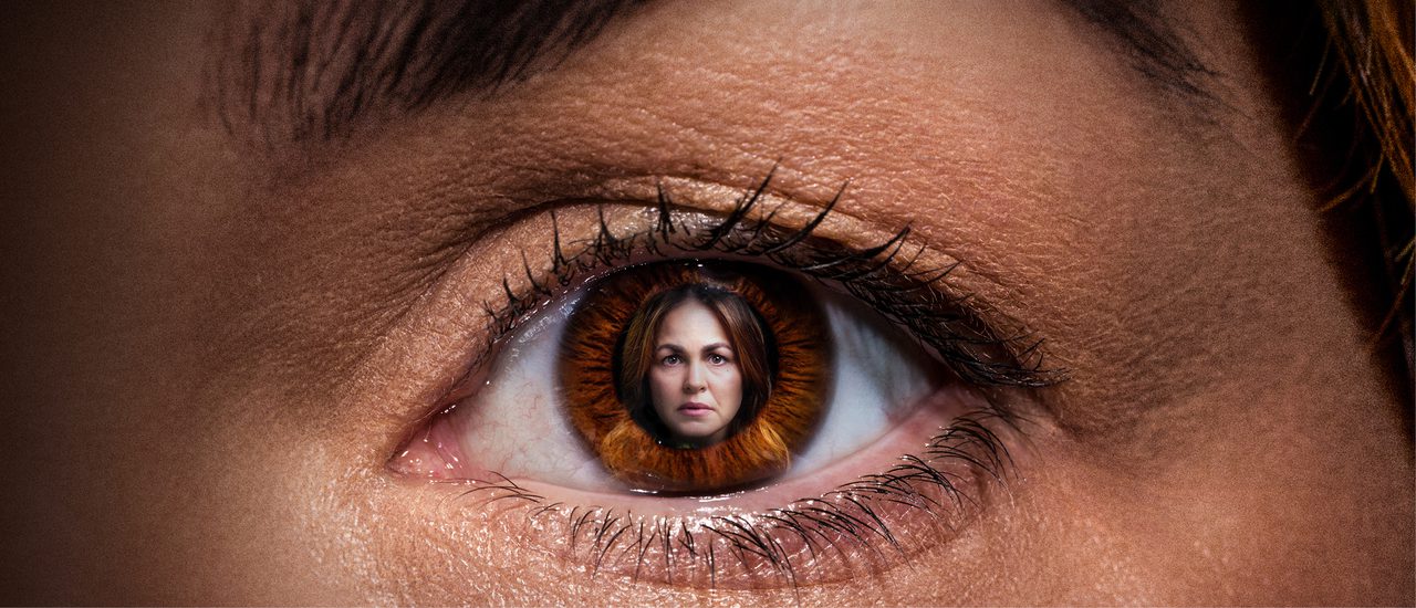 A close up of a woman's eye, within the pupil is an image of the same woman. She has dark brown hair and has a serious expression on her face.