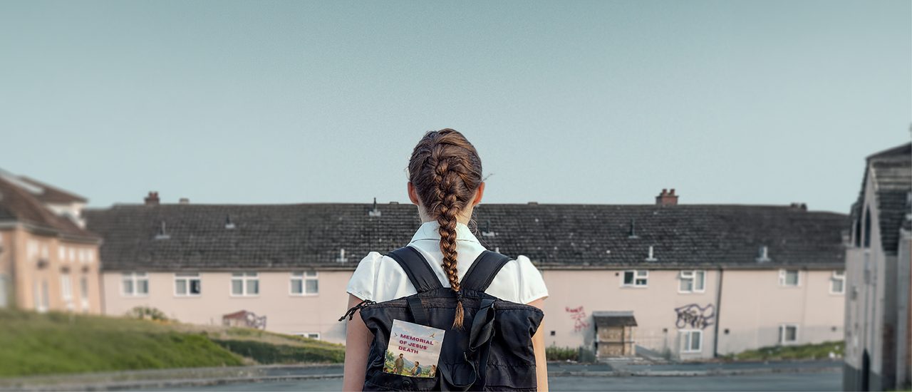 A young girl photographed from behind. She wears school uniform, a navy, pleated skirt and a short-sleeved, white blouse, and a black backpack. Her hair is tied back in a single, neat, French plait. A pamphlet which reads 'Memorial of Jesus' Death' can be seen poking out of her bag. A residential street with patches of grass, tarmac and terraced houses can be seen in the background, with a murky, grey sky.