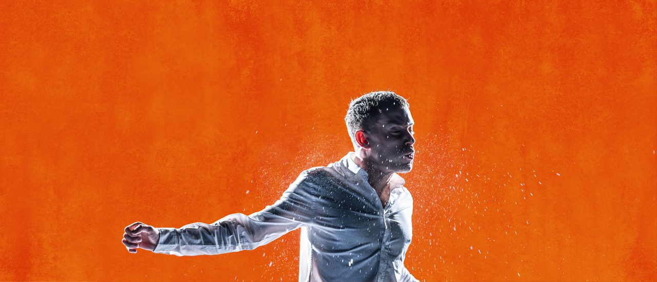 Against a bright orange background, a young man is in motion, as though mid-fight. He wears a white shirt. He holds one arm out to the side and his face looks pained. Water sprays around him.