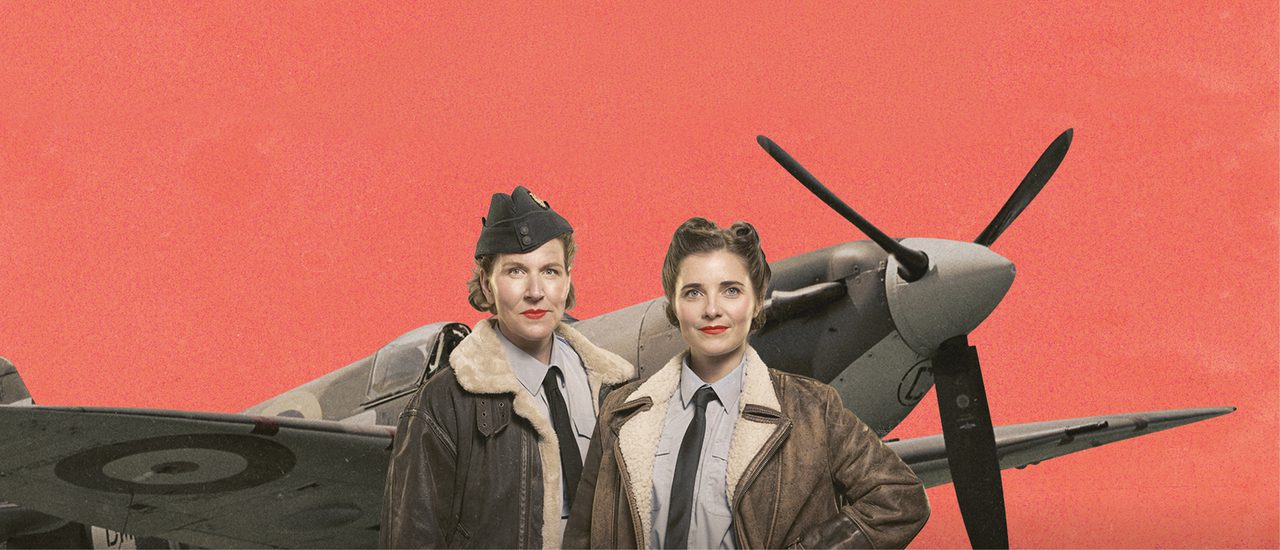 Two women in WWII RAF uniform, grey shirts, dark ties, brown aviator jackets and one in a dark hat, stand proudly together. They wear bright red lipstick and their hair is neatly styled in 1940s styles. They smile and look directly into the camera. Behind them is a spitfire plane against a coral background.