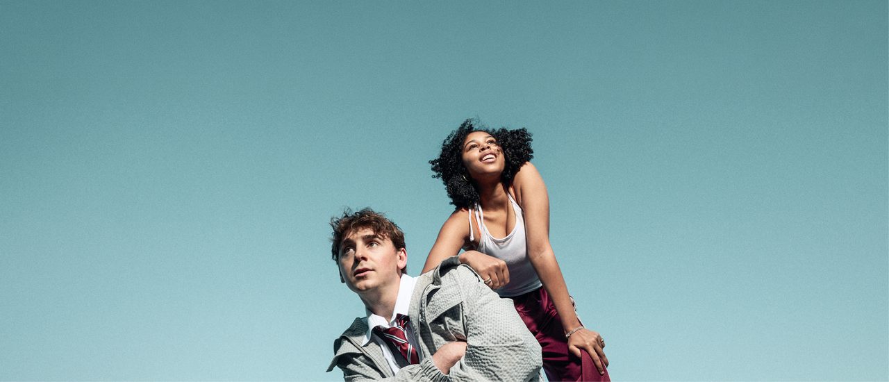 Two young people pose together against a light blue background. The boy crouches down, arms crossed over his chest, looking into the distance pensively. He wears school uniform and a thin bomber jacket. The girl stands, leaning on his shoulder with one arm and bending down with the other hand on her knee, looking up in the opposite direction with a smile. She wears burgundy, shiny tracksuit bottoms, a white vest top and hoop earrings.