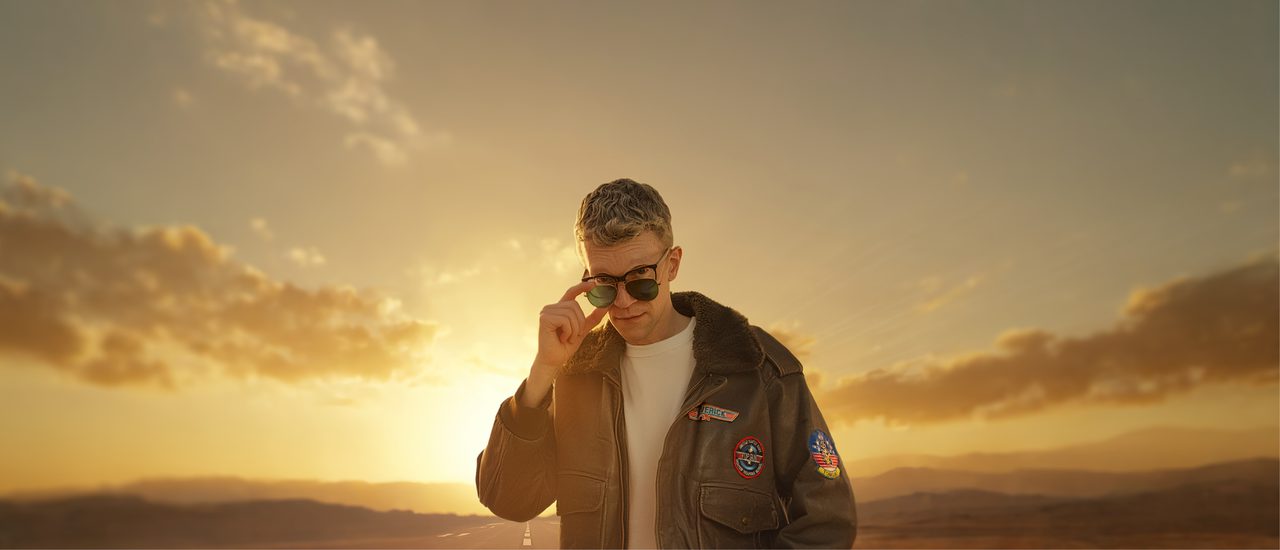 A spoof of an image from the iconic film Top Gun: a man stands wearing a leather aviator jacket, white t-shirt, blue jeans, glasses with thick, black frames, and aviator sunglasses. He holds the sunglasses lower down on the bridge of his nose and looks over them at the camera with an inquisitive facial expression. Behind him is a road and a dusty, golden sky.