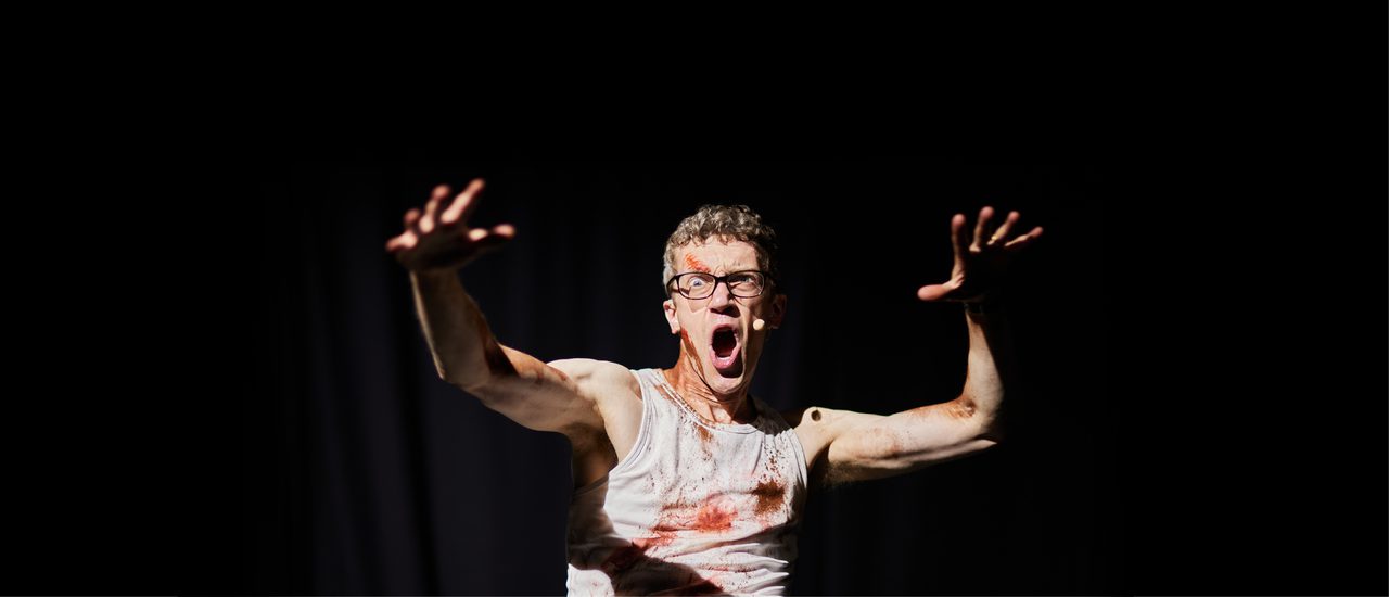Against a black curtain, a man stands in a dramatic pose: his hands are raised with his fingers splayed out and he appears to be shouting, with one eye wide. He wears glasses and a white vest and is smeared with blood.