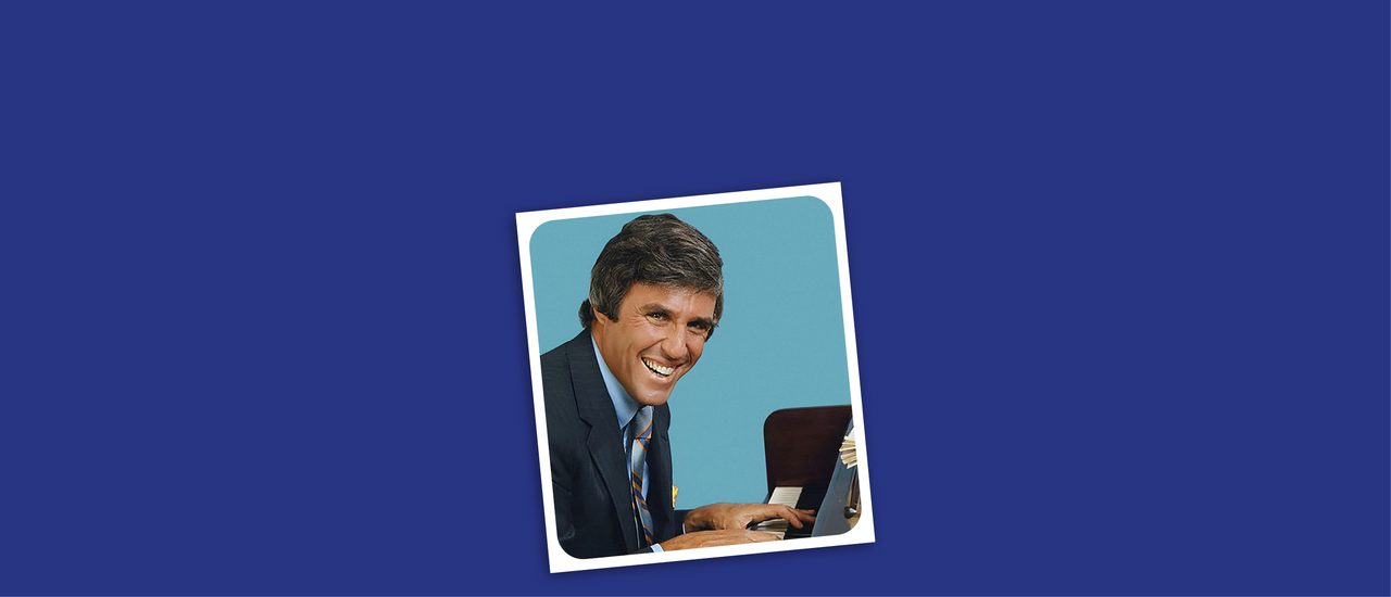A dark blue background with a tilted photo in the middle. In the photo, is a man in a suit sat at a piano. He is looking at the camera and smiling widely. Behind him is a light blue background with rounded corners and has a white border.
