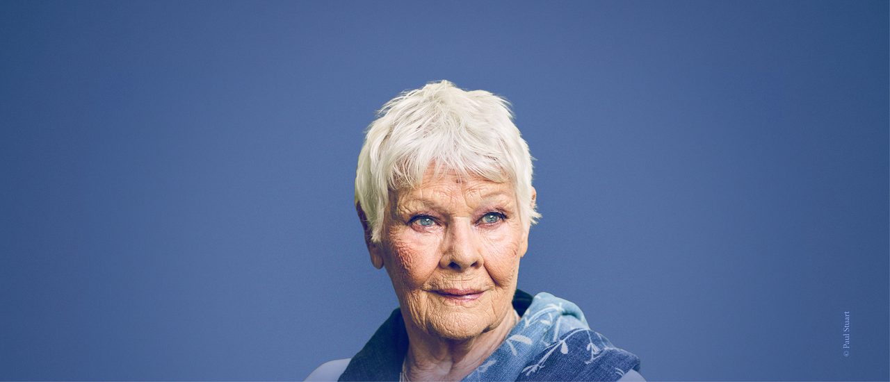 A photo of Dame Judi Dench on an indigo background. She looks off into the distance with a gentle smile. She has a blue scarf with white leaves draped over her shoulders.