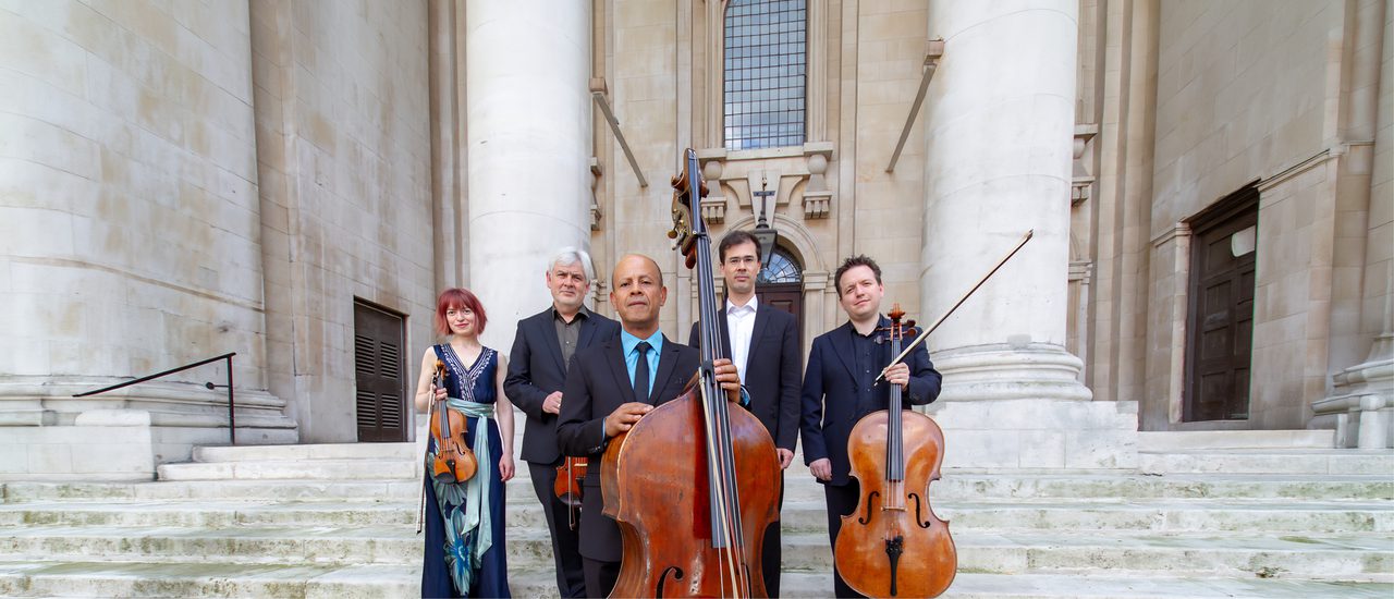 Five musicians holding different string instruments stand on the steps of an impressive sandstone building.
