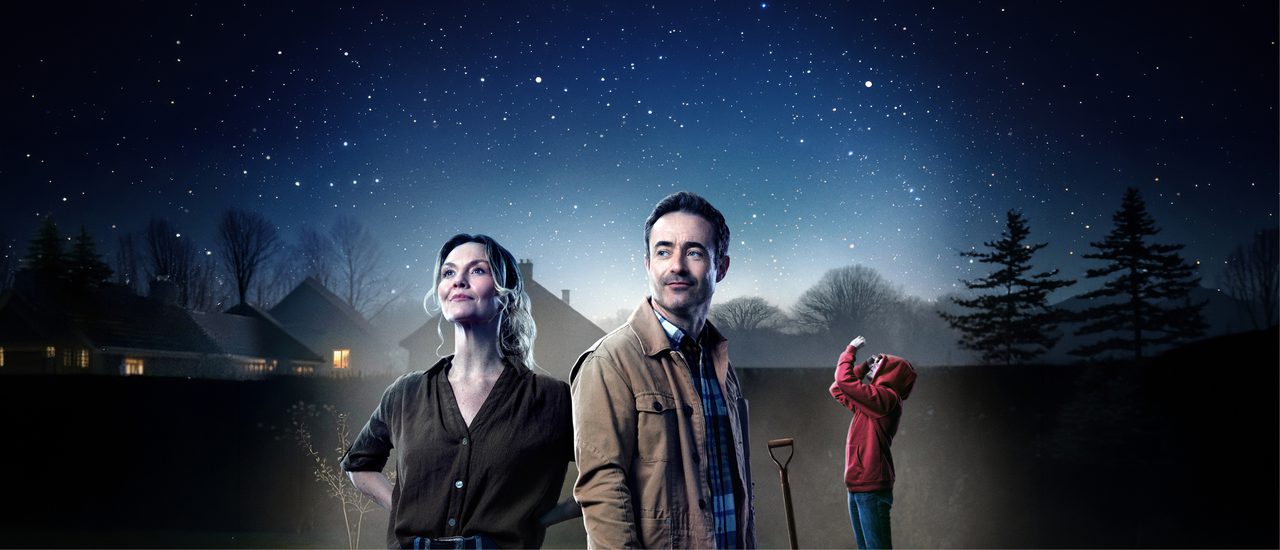 A casually dressed woman and man stand in a garden at night-time, against silhouetted houses, trees and a starry sky. Behind them, a young person in a red hoodie stands on an crime scene-type outline of a dog on the grass, with a pitchfork stuck into the ground.