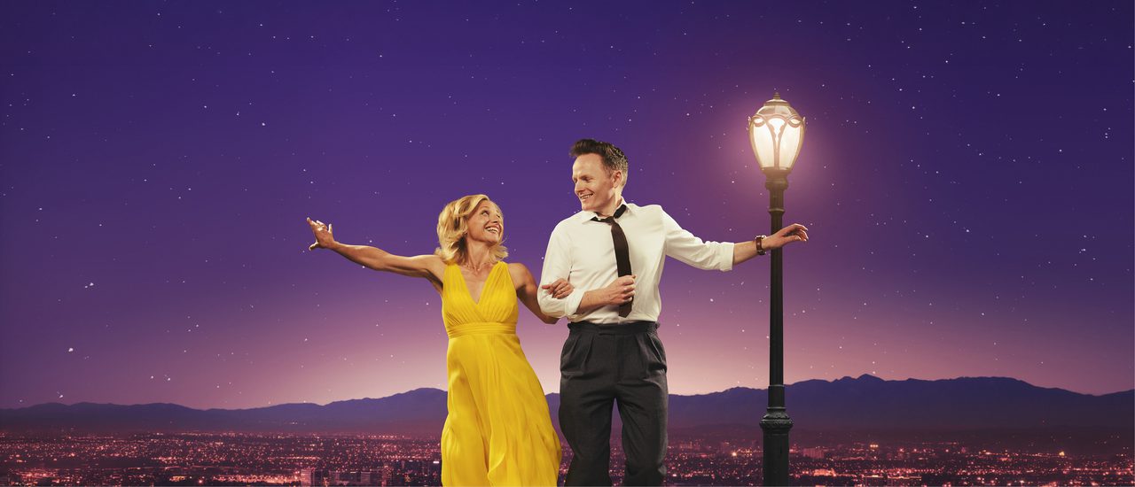 In a tribute to the film poster for La La Land, Liza Pulman and Joe Stilgoe dance together against a purple-hued vista of Hollywood, with a lighted street lamp beside them. She wears a sleeveless yellow dress, he is in white shirt, black tie and trousers, and they smile happily at each other as they leap off the ground.